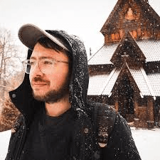 Bearded man in glasses and hood stands in falling snow before a wooden stave church.